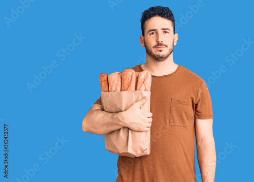 Young hispanic man holding paper bag with bread thinking attitude and sober expression looking self confident photo