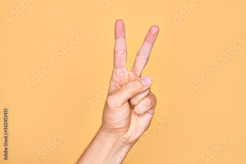 Hand of caucasian young man showing fingers over isolated yellow background counting number 2 showing two fingers, gesturing victory and winner symbol