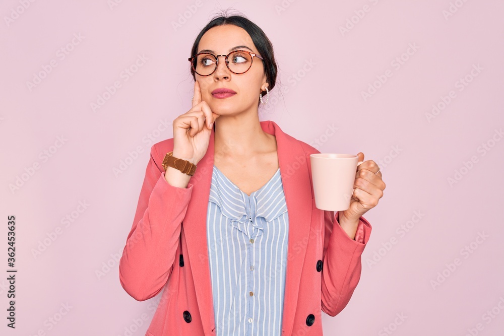 Young beautiful businesswoman with blue eyes wearing glasses drinking pink mug of coffee serious face thinking about question, very confused idea Young beautiful businesswoman with blue eyes wearing glasses drinking pink mug of coffee serious face thinking about question, very confused idea