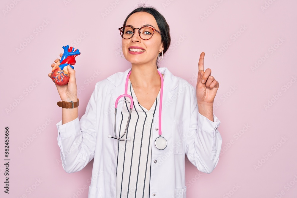 Young beautiful cardiologist woman with blue eyes wearing stethoscope holding heart surprised with an idea or question pointing finger with happy face, number one Young beautiful cardiologist woman with blue eyes wearing stethoscope holding heart surprised with an idea or question pointing finger with happy face, number one