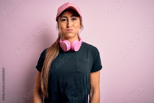 Young beautiful fitness sports woman wearing training cap and headphones over pink background puffing cheeks with funny face. Mouth inflated with air, crazy expression.