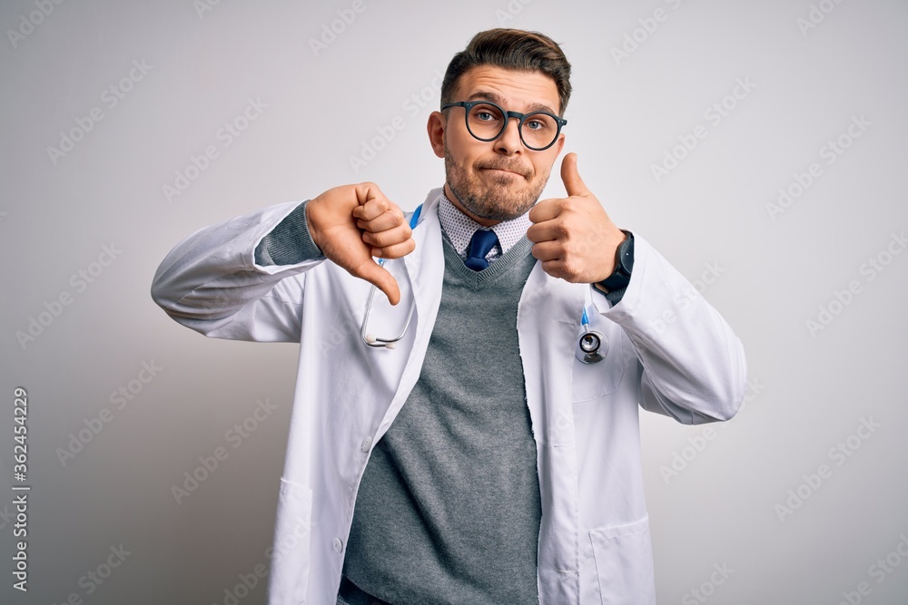 Young doctor man with blue eyes wearing medical coat and stethoscope over isolated background Doing thumbs up and down, disagreement and agreement expression. Crazy conflict Young doctor man with blue eyes wearing medical coat and stethoscope over isolated background Doing thumbs up and down, disagreement and agreement expression. Crazy conflict