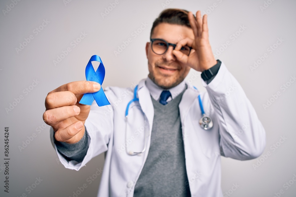 Young doctor man wearing medical coat and holding colon cancer awareness blue ribbon with happy face smiling doing ok sign with hand on eye looking through fingers Young doctor man wearing medical coat and holding colon cancer awareness blue ribbon with happy face smiling doing ok sign with hand on eye looking through fingers