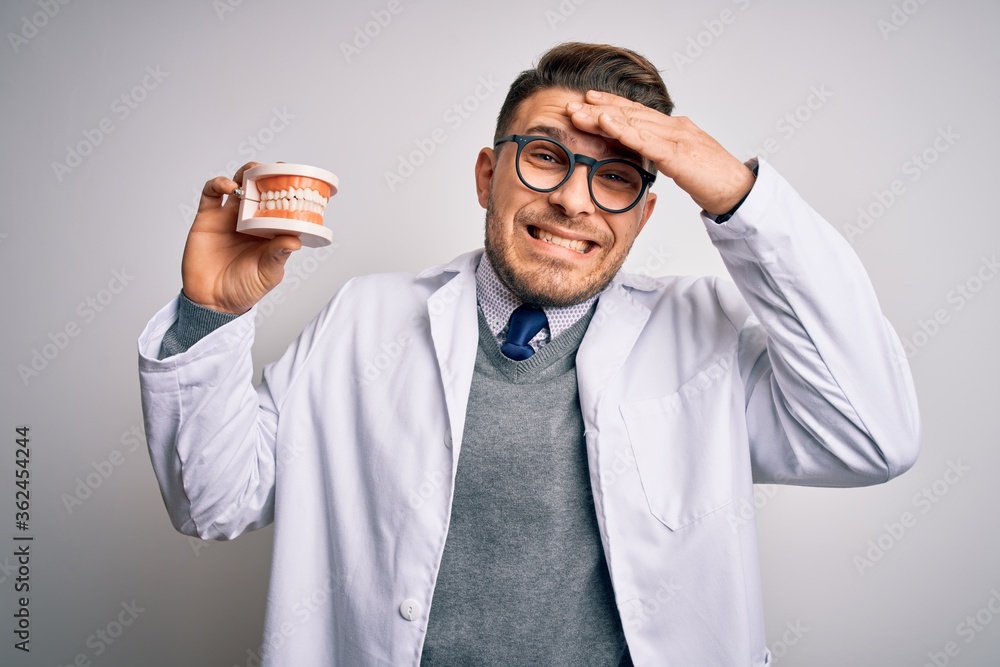 Young dentist man with blue eyes holding orthodontic dental prosthesis over isolated background stressed with hand on head, shocked with shame and surprise face, angry and frustrated. Fear and upset. Young dentist man with blue eyes holding orthodontic dental prosthesis over isolated background stressed with hand on head, shocked with shame and surprise face, angry and frustrated. Fear and upset.