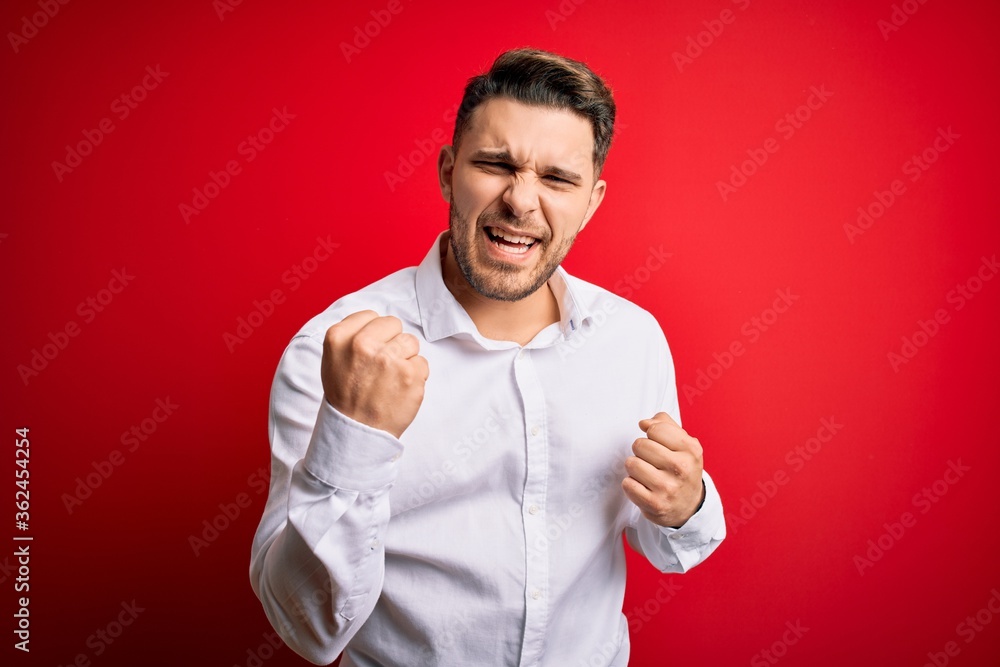 Young business man with blue eyes wearing elegant shirt standing over red isolated background celebrating surprised and amazed for success with arms raised and eyes closed. Winner concept. Young business man with blue eyes wearing elegant shirt standing over red isolated background celebrating surprised and amazed for success with arms raised and eyes closed. Winner concept.
