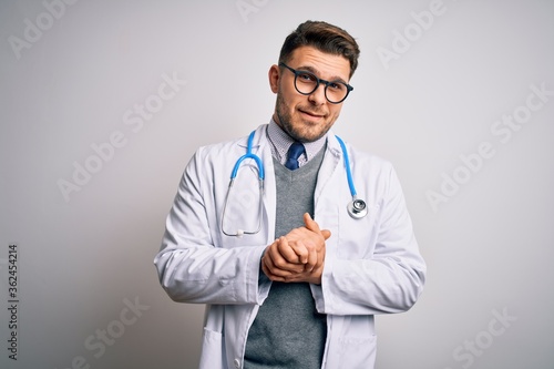 Young doctor man with blue eyes wearing medical coat and stethoscope over isolated background with hands together and crossed fingers smiling relaxed and cheerful. Success and optimistic