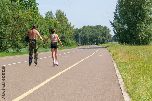 People are resting in the park. A man and a teenager girl rollerblading hold hands. View from the back. Road marking  green trees. Concept - family vacation   fathers day.