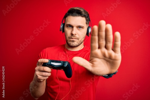 Young gamer man with blue eyes playing video games using gamepad joystick with open hand doing stop sign with serious and confident expression, defense gesture