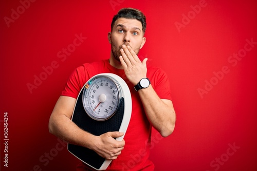 Young fitness man with blue eyes holding scale dieting for healthy weight over red background cover mouth with hand shocked with shame for mistake, expression of fear, scared in silence.