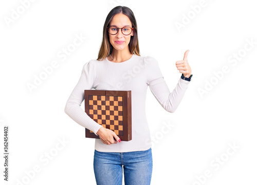 Beautiful brunette young woman holding chess smiling happy and positive, thumb up doing excellent and approval sign