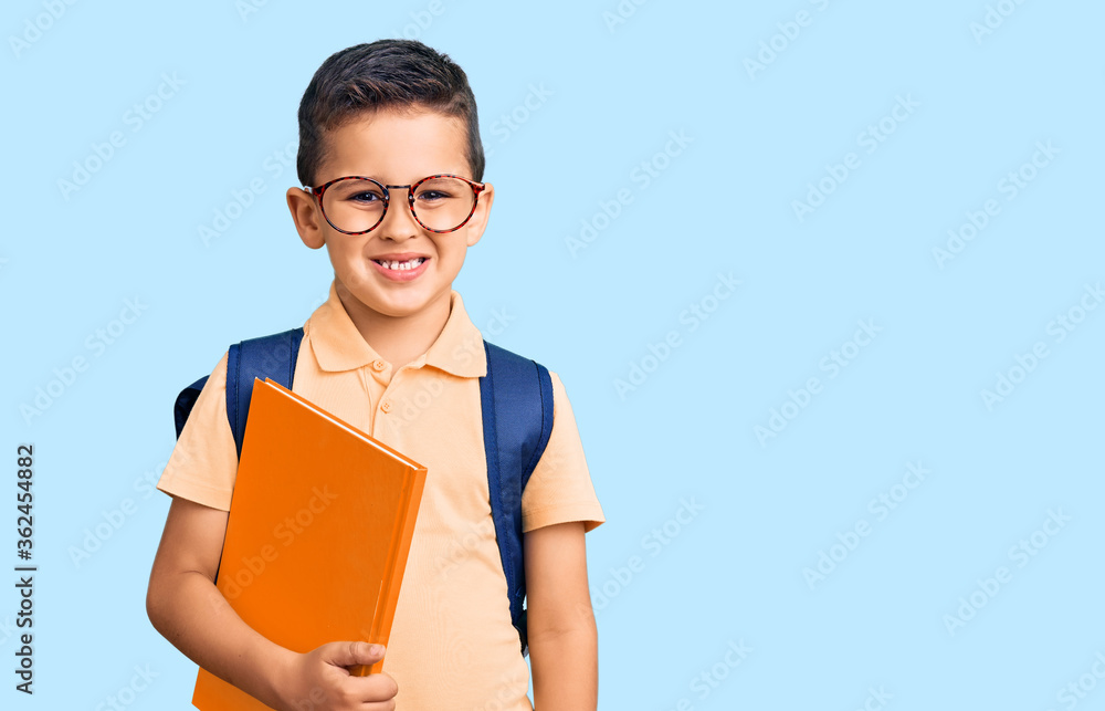 Obraz premium Little cute boy kid wearing school bag and holding book looking positive and happy standing and smiling with a confident smile showing teeth