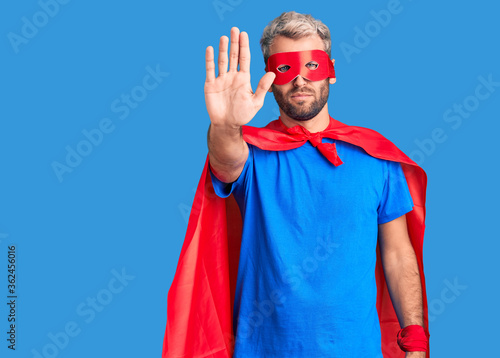 Young blond man wearing super hero custome doing stop sing with palm of the hand. warning expression with negative and serious gesture on the face.
