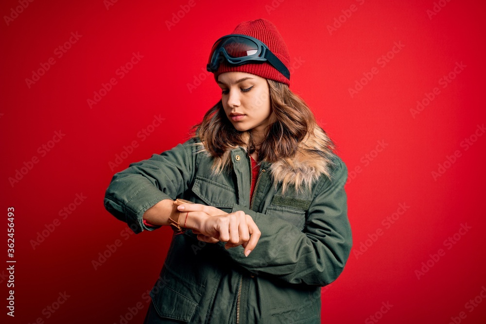 Young blonde girl wearing ski glasses and winter coat for ski weather over red background Checking the time on wrist watch, relaxed and confident Young blonde girl wearing ski glasses and winter coat for ski weather over red background Checking the time on wrist watch, relaxed and confident