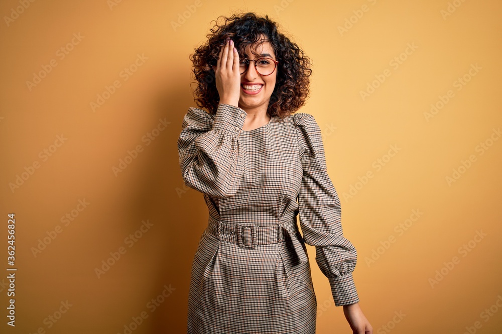 Beautiful arab business woman wearing dress and glasses standing over yellow background covering one eye with hand, confident smile on face and surprise emotion. Beautiful arab business woman wearing dress and glasses standing over yellow background covering one eye with hand, confident smile on face and surprise emotion.