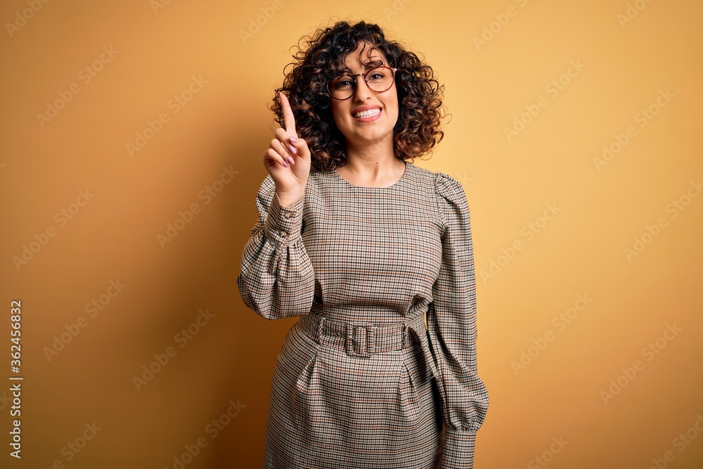 Beautiful arab business woman wearing dress and glasses standing over yellow background pointing finger up with successful idea. Exited and happy. Number one. Beautiful arab business woman wearing dress and glasses standing over yellow background pointing finger up with successful idea. Exited and happy. Number one.