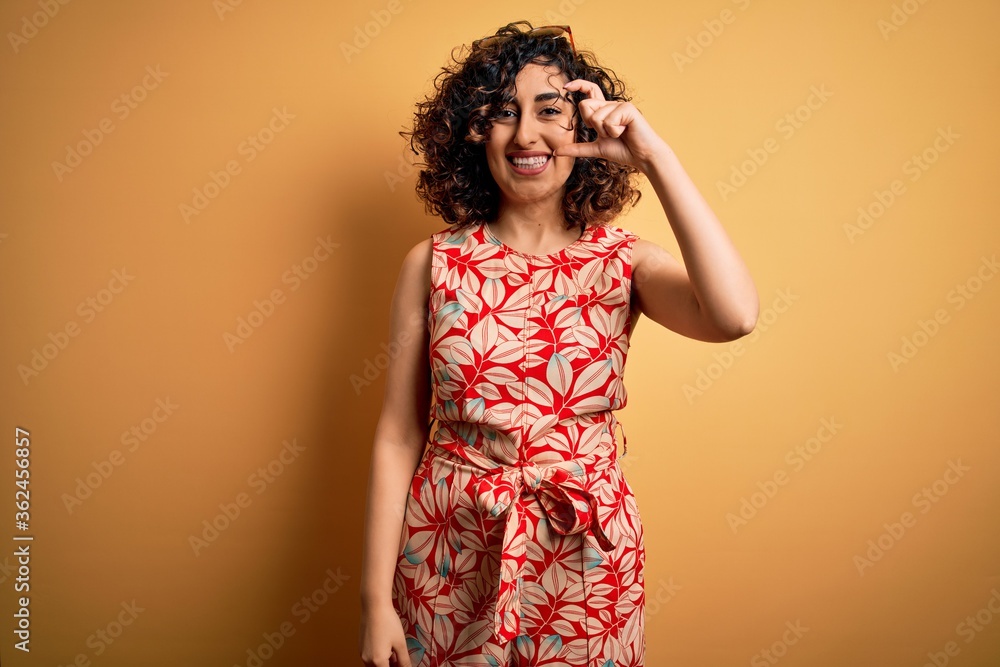 Young beautiful curly arab woman on vacation wearing summer floral dress and sunglasses smiling and confident gesturing with hand doing small size sign with fingers looking and the camera. Measure. Young beautiful curly arab woman on vacation wearing summer floral dress and sunglasses smiling and confident gesturing with hand doing small size sign with fingers looking and the camera. Measure.