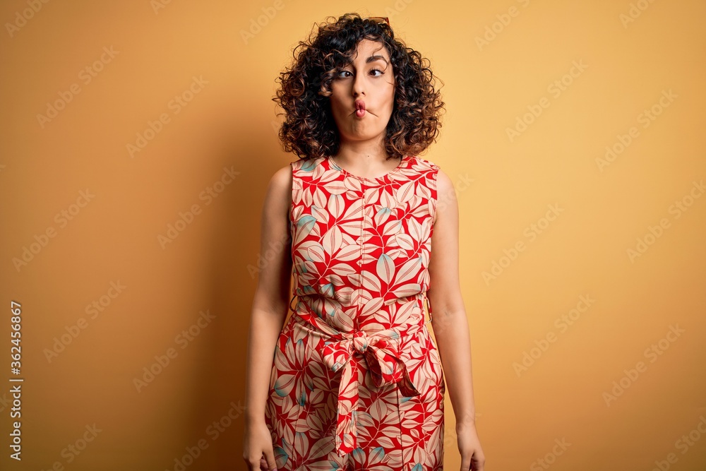 Young beautiful curly arab woman on vacation wearing summer floral dress and sunglasses making fish face with lips, crazy and comical gesture. Funny expression. Young beautiful curly arab woman on vacation wearing summer floral dress and sunglasses making fish face with lips, crazy and comical gesture. Funny expression.