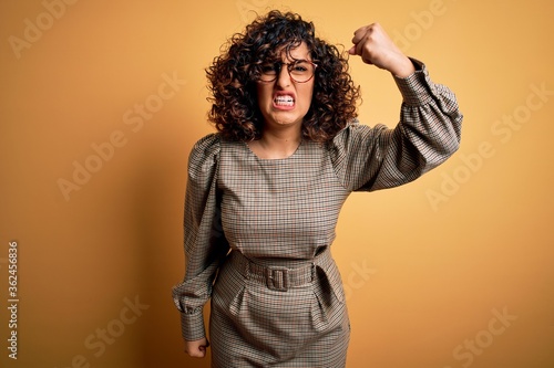 Beautiful arab business woman wearing dress and glasses standing over yellow background angry and mad raising fist frustrated and furious while shouting with anger. Rage and aggressive concept. © Krakenimages.com