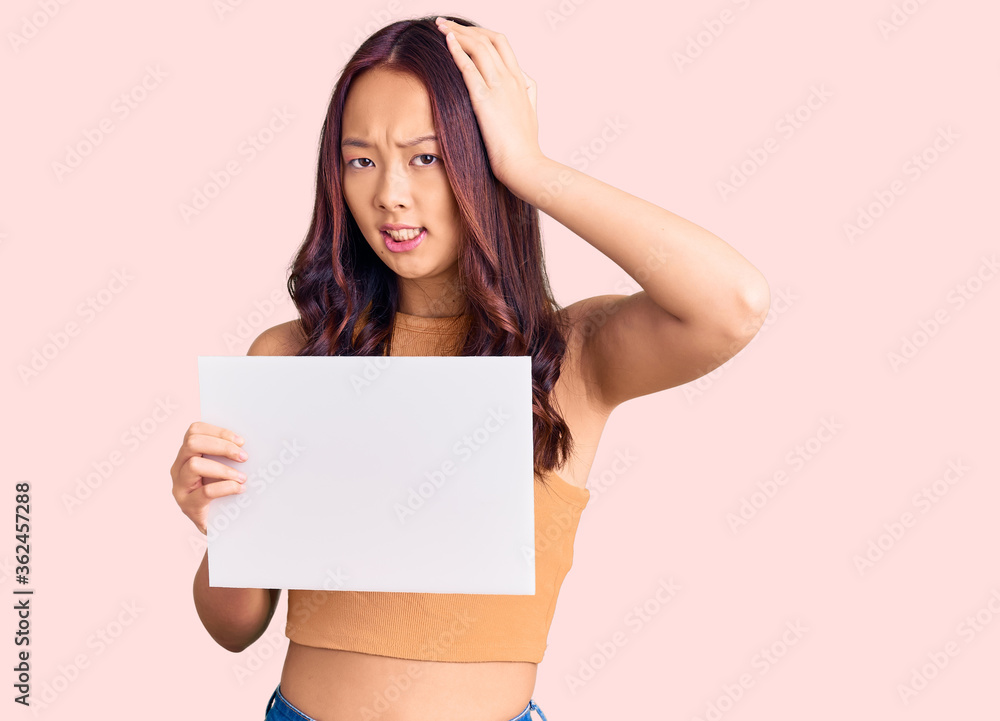 Young beautiful chinese girl holding paper banner with blank space stressed and frustrated with hand on head, surprised and angry face Young beautiful chinese girl holding paper banner with blank space stressed and frustrated with hand on head, surprised and angry face