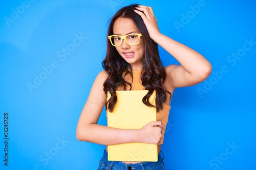 Young beautiful chinese girl wearing glasses holding book stressed and frustrated with hand on head, surprised and angry face