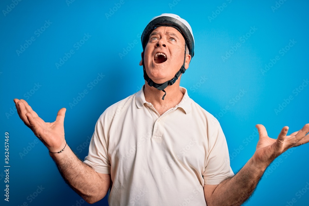 Naklejka premium Middle age hoary cyclist man wearing bike security helmet over isolated blue background crazy and mad shouting and yelling with aggressive expression and arms raised. Frustration concept.