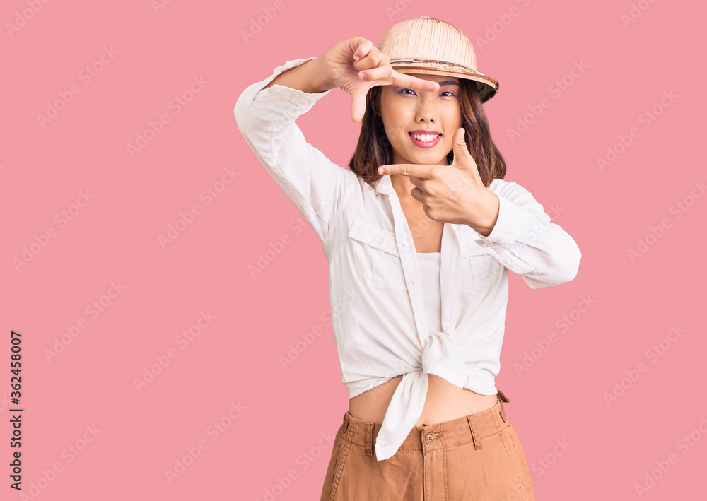 Young beautiful chinese girl wearing explorer hat smiling making frame with hands and fingers with happy face. creativity and photography concept. Young beautiful chinese girl wearing explorer hat smiling making frame with hands and fingers with happy face. creativity and photography concept.