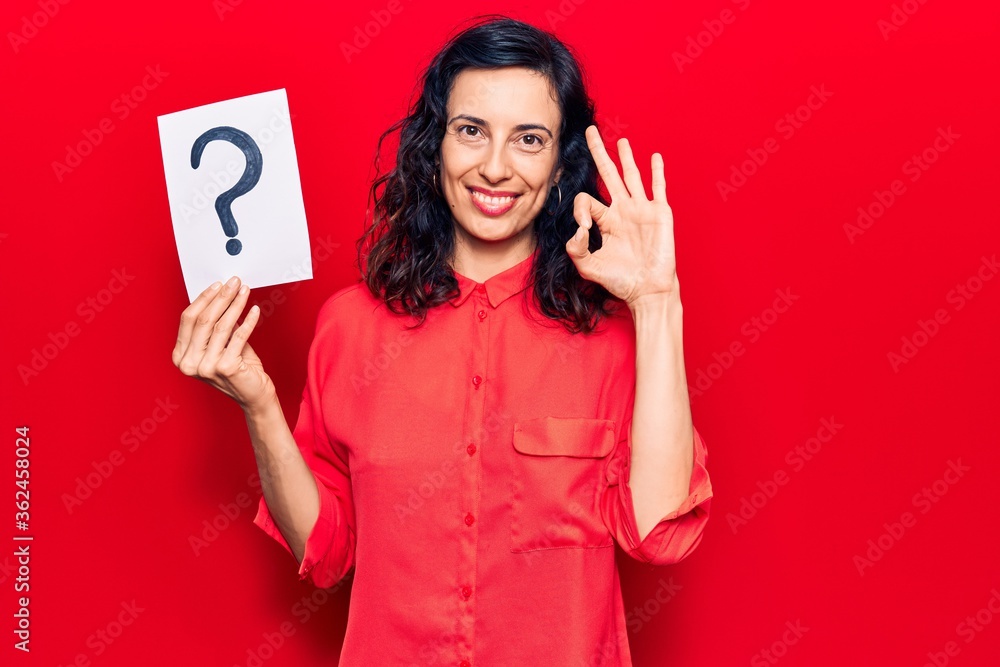Naklejka premium Young beautiful hispanic woman holding question mark doing ok sign with fingers, smiling friendly gesturing excellent symbol