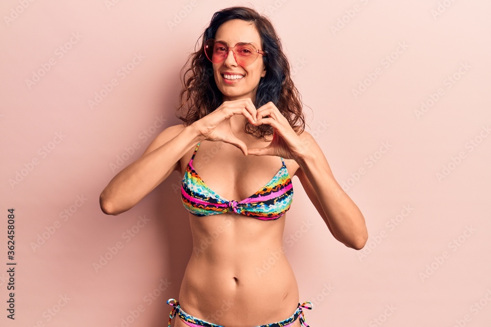 Naklejka premium Young beautiful hispanic woman wearing bikini and heart sunglasses smiling in love doing heart symbol shape with hands. romantic concept.