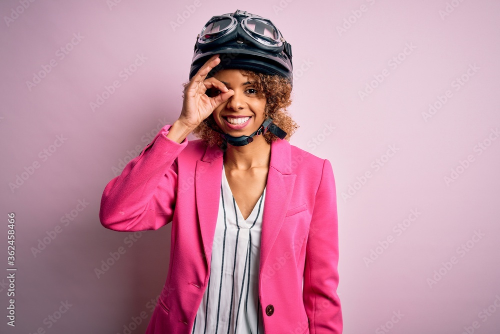 African american motorcyclist woman with curly hair wearing moto helmet over pink background doing ok gesture with hand smiling, eye looking through fingers with happy face. African american motorcyclist woman with curly hair wearing moto helmet over pink background doing ok gesture with hand smiling, eye looking through fingers with happy face.