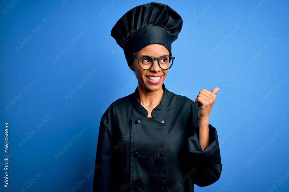 Young african american chef woman wearing cooker uniform and hat over blue background smiling with happy face looking and pointing to the side with thumb up. Young african american chef woman wearing cooker uniform and hat over blue background smiling with happy face looking and pointing to the side with thumb up.