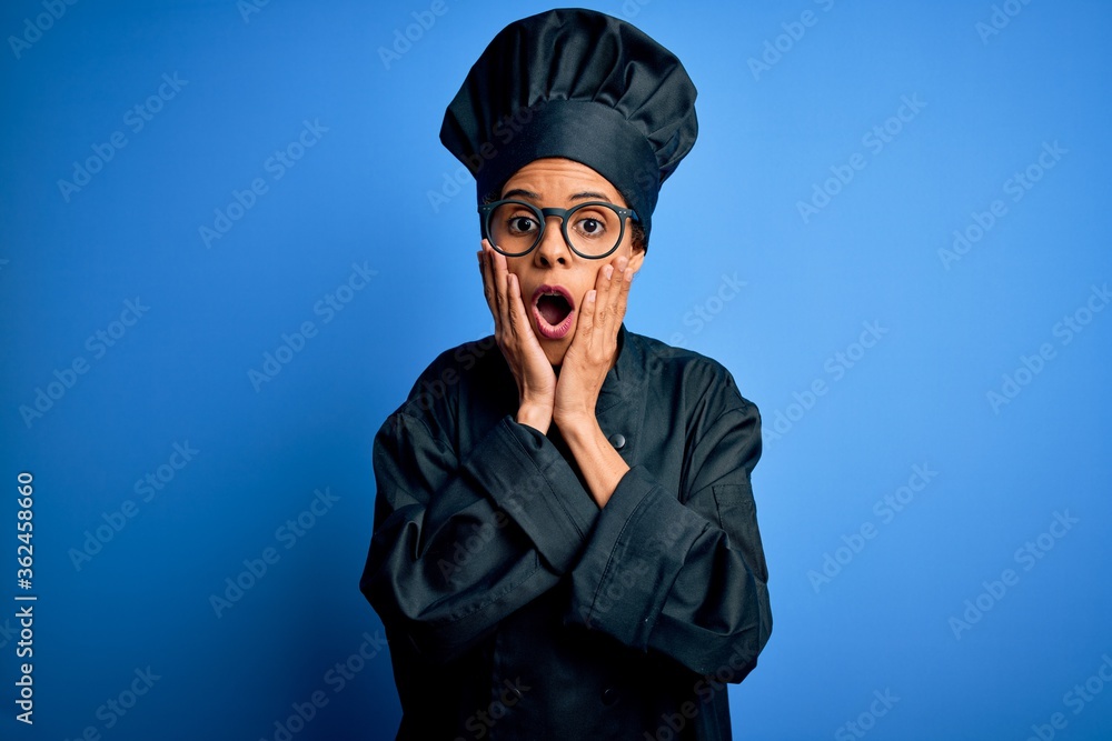Young african american chef woman wearing cooker uniform and hat over blue background afraid and shocked, surprise and amazed expression with hands on face Young african american chef woman wearing cooker uniform and hat over blue background afraid and shocked, surprise and amazed expression with hands on face