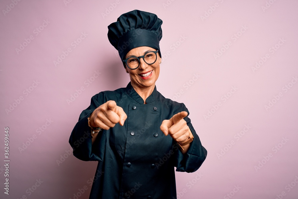 Middle age brunette chef woman wearing cooker uniform and hat over isolated pink background pointing to you and the camera with fingers, smiling positive and cheerful Middle age brunette chef woman wearing cooker uniform and hat over isolated pink background pointing to you and the camera with fingers, smiling positive and cheerful