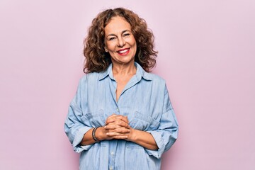 Middle age beautiful woman wearing casual denim shirt standing over pink background with hands together and crossed fingers smiling relaxed and cheerful. Success and optimistic