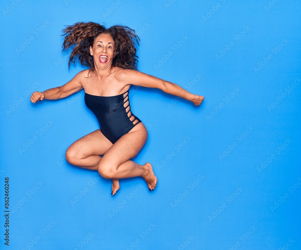 Middle age beautiful hispanic woman on vacation wearing swimsuit smiling happy. Jumping with smile on face celebrating over isolated blue background Middle age beautiful hispanic woman on vacation wearing swimsuit smiling happy. Jumping with smile on face celebrating over isolated blue background