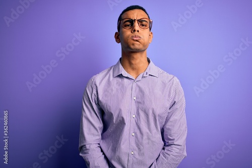 Handsome african american man wearing striped shirt and glasses over purple background puffing cheeks with funny face. Mouth inflated with air, crazy expression. © Krakenimages.com