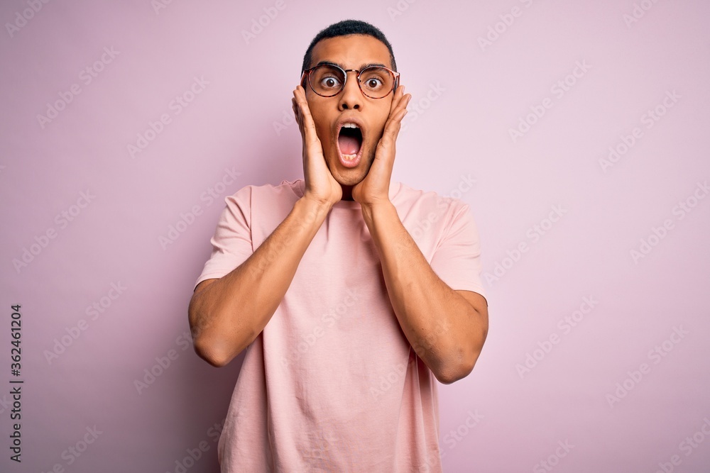 Handsome african american man wearing casual t-shirt and glasses over pink background afraid and shocked, surprise and amazed expression with hands on face Handsome african american man wearing casual t-shirt and glasses over pink background afraid and shocked, surprise and amazed expression with hands on face