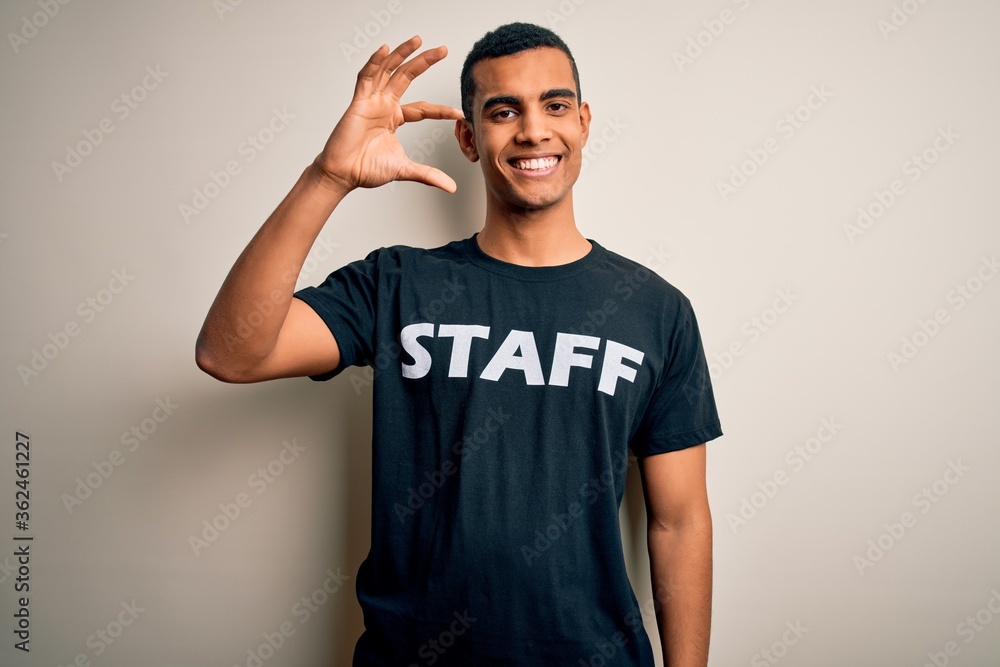 Young handsome african american worker man wearing staff uniform over white background smiling and confident gesturing with hand doing small size sign with fingers looking and the camera. Measure. Young handsome african american worker man wearing staff uniform over white background smiling and confident gesturing with hand doing small size sign with fingers looking and the camera. Measure.