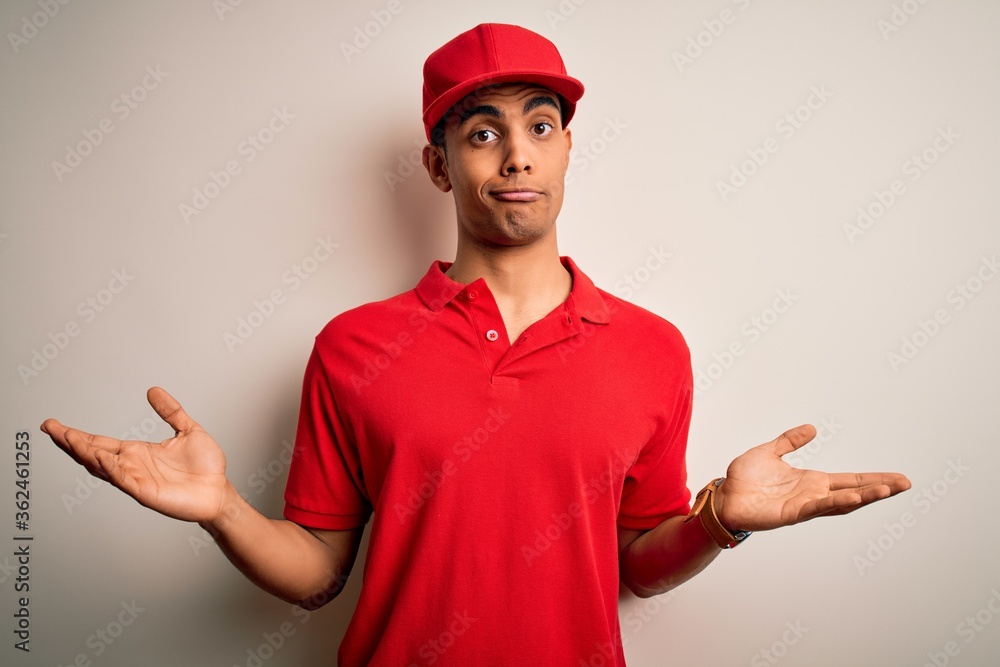 Young handsome african american man wearing casual polo and cap over red background clueless and confused expression with arms and hands raised. Doubt concept. Young handsome african american man wearing casual polo and cap over red background clueless and confused expression with arms and hands raised. Doubt concept.