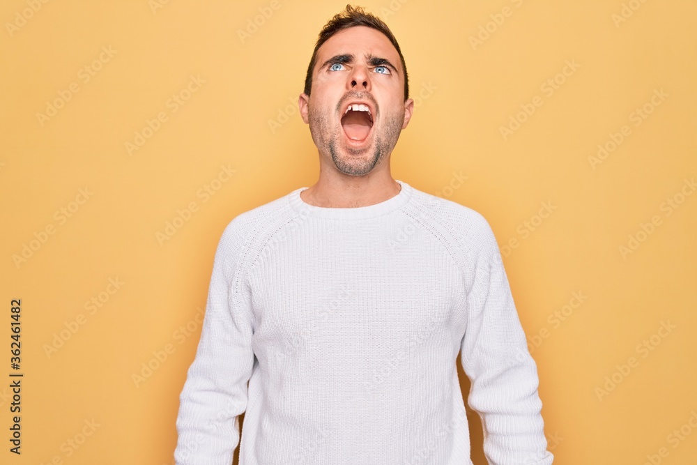 Young handsome man with blue eyes wearing casual sweater standing over yellow background angry and mad screaming frustrated and furious, shouting with anger. Rage and aggressive concept. Young handsome man with blue eyes wearing casual sweater standing over yellow background angry and mad screaming frustrated and furious, shouting with anger. Rage and aggressive concept.
