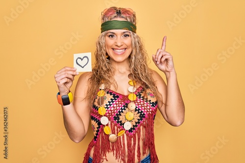 Beautiful blonde hippie woman wearing sunglasses and accessories holding reminder with heart surprised with an idea or question pointing finger with happy face, number one