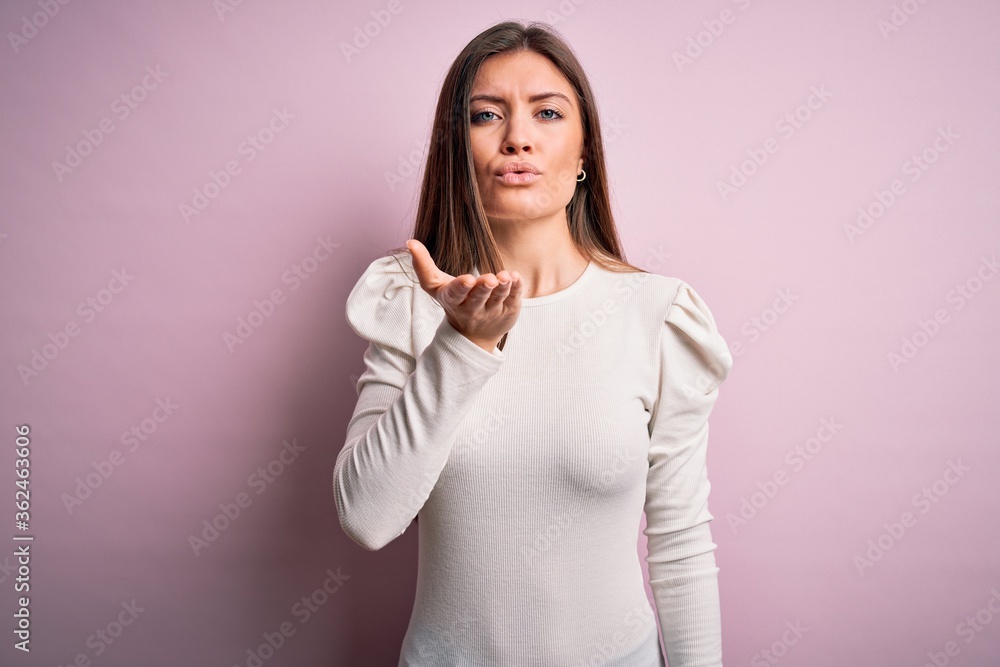 Young beautiful woman with blue eyes wearing casual white t-shirt over pink background looking at the camera blowing a kiss with hand on air being lovely and sexy. Love expression. Young beautiful woman with blue eyes wearing casual white t-shirt over pink background looking at the camera blowing a kiss with hand on air being lovely and sexy. Love expression.