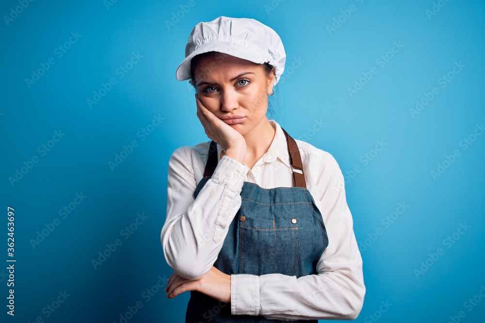 Young beautiful baker woman with blue eyes wearing apron and cap over blue background thinking looking tired and bored with depression problems with crossed arms. Young beautiful baker woman with blue eyes wearing apron and cap over blue background thinking looking tired and bored with depression problems with crossed arms.