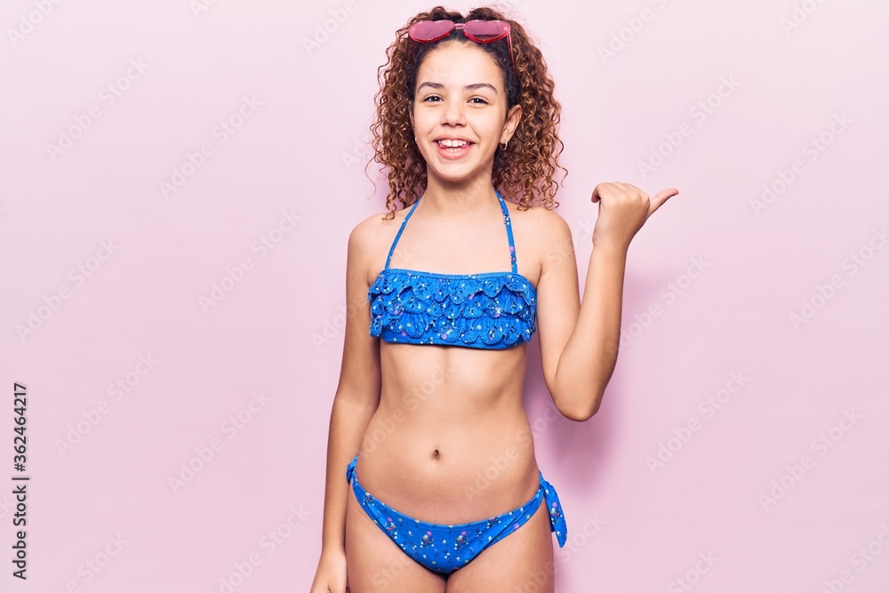 Naklejka premium Beautiful kid girl with curly hair wearing bikini and sunglasses smiling with happy face looking and pointing to the side with thumb up.