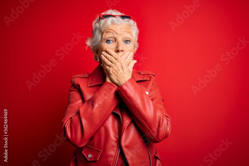 Senior beautiful grey-haired woman wearing casual red jacket and sunglasses shocked covering mouth with hands for mistake. Secret concept.