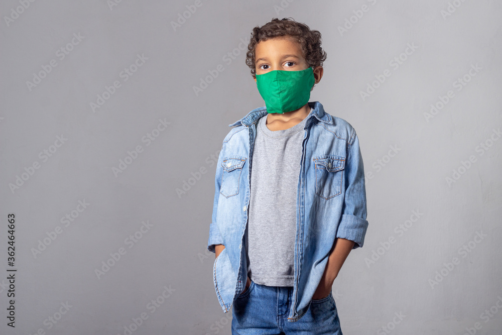 White curly-haired boy wearing protective mask against covid-19 wearing gray t-shirt overlaid by denim shirt on gray background White curly-haired boy wearing protective mask against covid-19 wearing gray t-shirt overlaid by denim shirt on gray background
