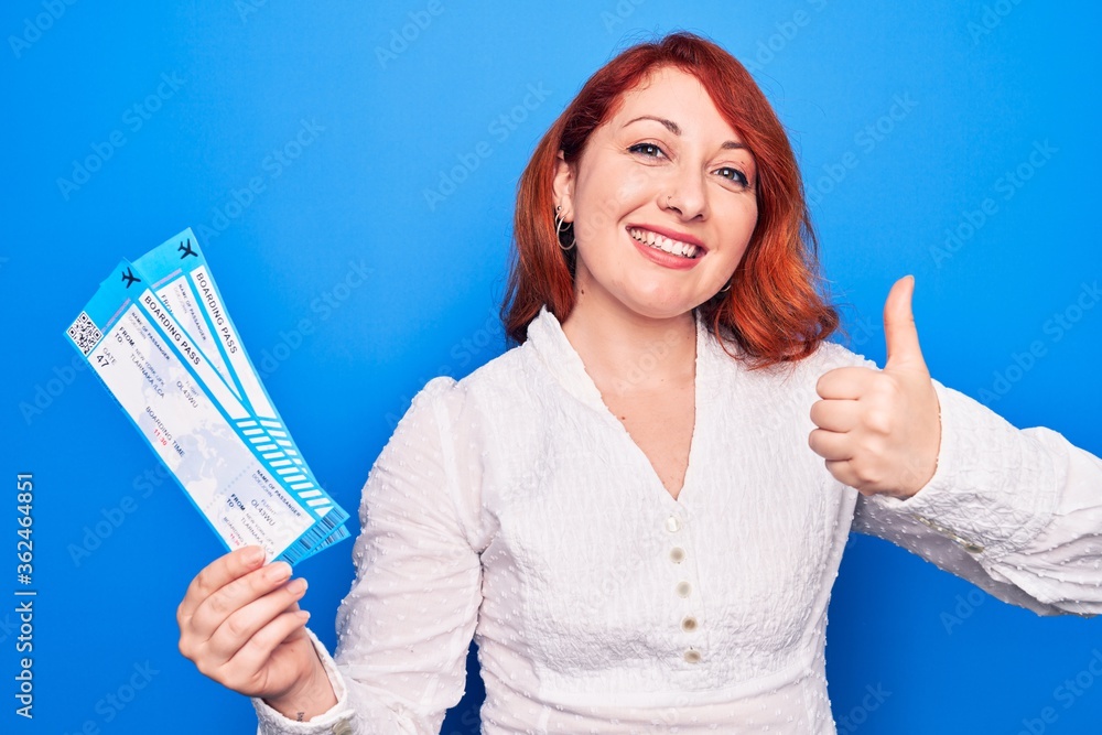 Young redhead tourist woman on vacation holding airline boarding pass over blue background smiling happy and positive, thumb up doing excellent and approval sign Young redhead tourist woman on vacation holding airline boarding pass over blue background smiling happy and positive, thumb up doing excellent and approval sign