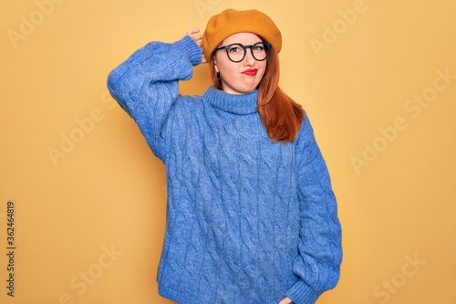Young beautiful redhead woman wearing french beret and glasses over yellow background confuse and wonder about question. Uncertain with doubt, thinking with hand on head. Pensive concept. © Krakenimages.com