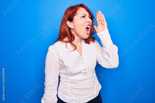 Young beautiful redhead businesswoman wearing elegant shirt standing over blue background shouting and screaming loud to side with hand on mouth. Communication concept.