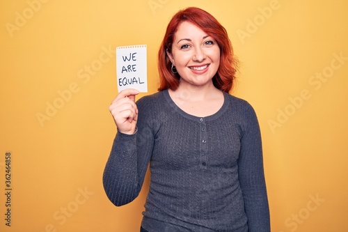 Young beautiful redhead woman asking for equality holding paper with we are equal message looking positive and happy standing and smiling with a confident smile showing teeth