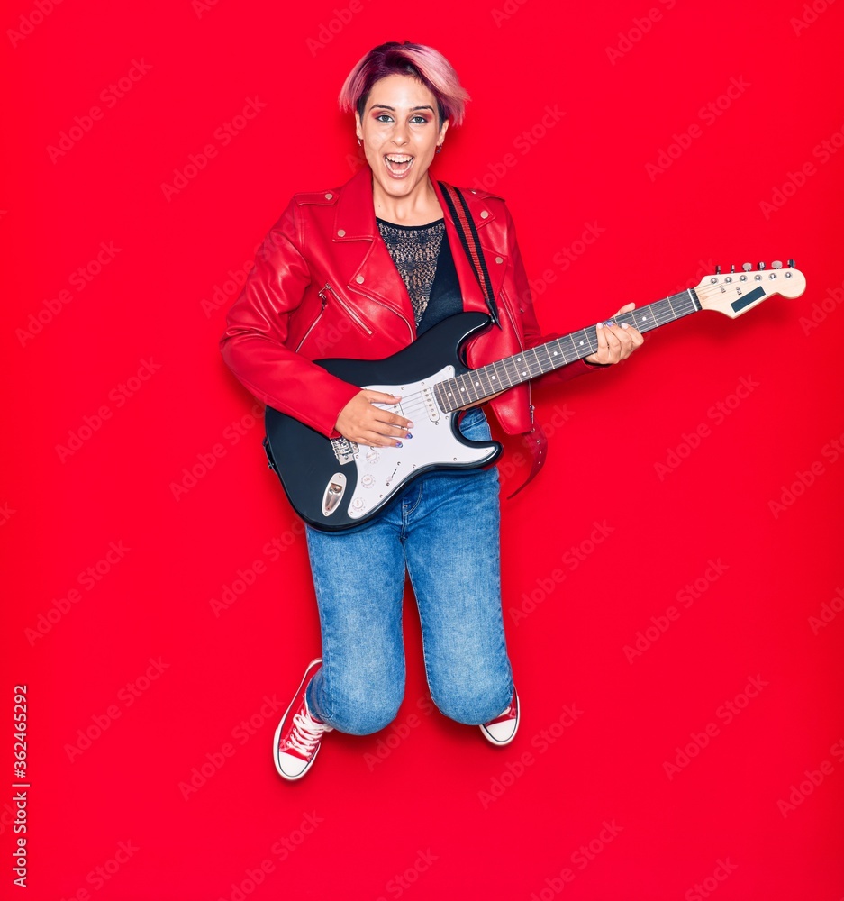 Young beautiful guitarist woman with pink short hair playing electric guitar smiling happy. Jumping with smile on face over isolated red background Young beautiful guitarist woman with pink short hair playing electric guitar smiling happy. Jumping with smile on face over isolated red background